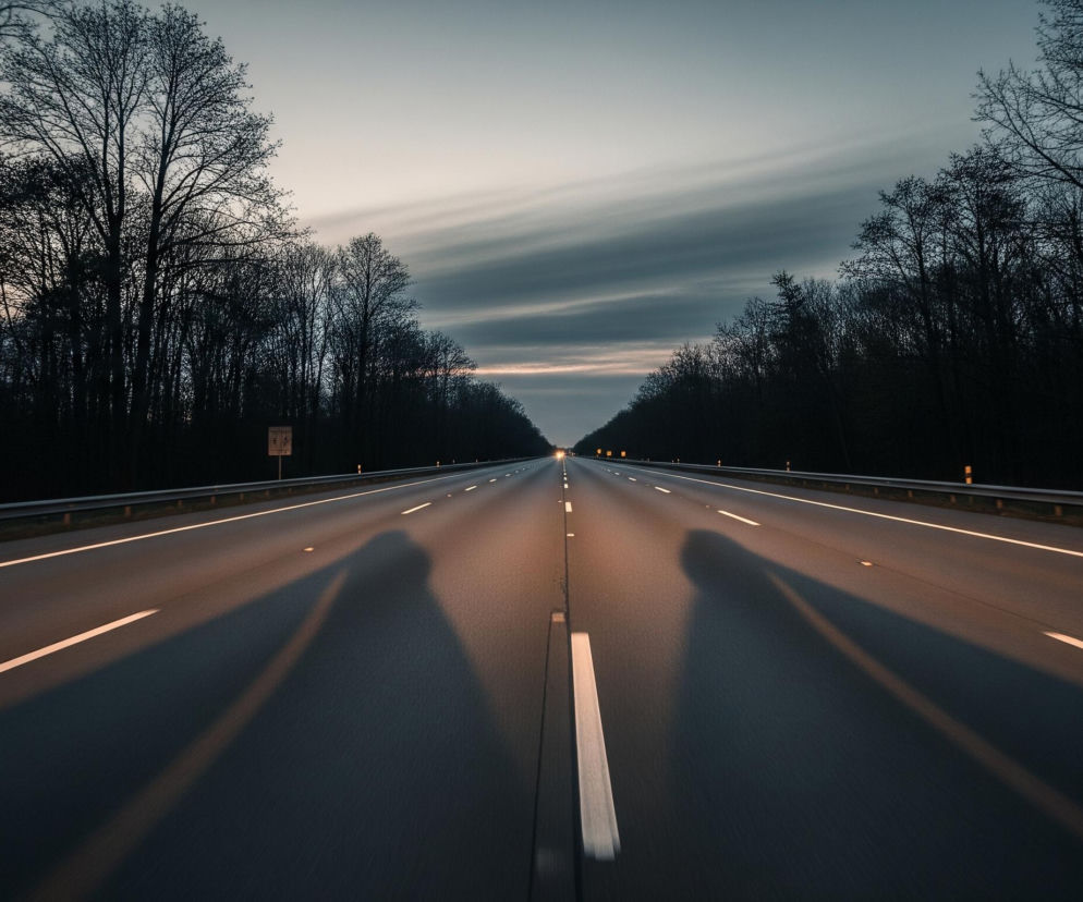 Długa, prosta droga biegnie w głąb kadru, otoczona po obu stronach ciemnymi, nagimi drzewami, tworzącymi gęsty las. Nawierzchnia asfaltowa ma kolor ciemnoszary, z wyraźnymi białymi liniami przerywanymi i ciągłymi, wyznaczającymi pasy ruchu oraz krawędzie jezdni. Na drodze widoczne są długie, rozciągnięte cienie, prawdopodobnie pochodzące od obiektu fotografującego, rzucane w kierunku przeciwnym do oddalającego się światła na horyzoncie. Niebo nad lasem, szczególnie w centralnej części, rozjaśnia się delikatnymi odcieniami pomarańczu i różu, przechodzącymi w szare, pofalowane chmury, które bledną ku górze kadru, gdzie dominuje jaśniejszy błękit. W oddali na drodze widoczne są jaskrawe, punktowe światła, wskazujące na obecność innych pojazdów lub oświetlenia drogowego, podkreślając perspektywę i oddalenie.