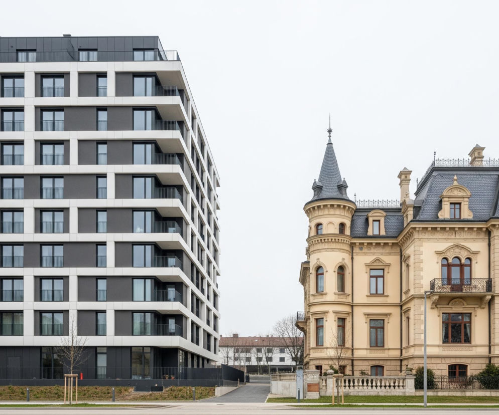 Na obrazie widoczne są dwa budynki o kontrastującej architekturze. Po lewej stronie dominuje nowoczesny, wielopiętrowy budynek o szarej elewacji z geometrycznymi, białymi ramami, które tworzą balkony z czarnymi balustradami. Po prawej stronie stoi klasyczna willa o beżowych ścianach, bogato zdobiona detalami architektonicznymi, z wieżyczką i ciemnoszarym spadzistym dachem. Pomiędzy budynkami znajduje się utwardzony obszar z pojedynczymi, młodymi drzewkami i trawą, a w tle widać fragmenty innych budynków oraz nagie drzewa.