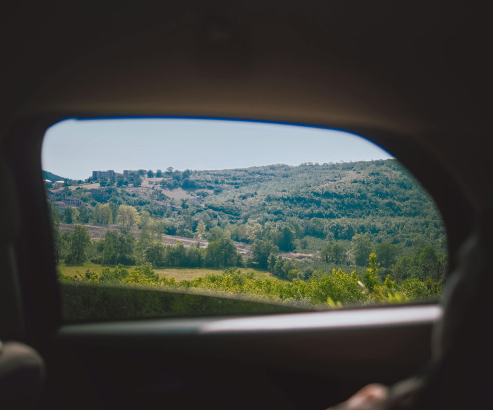 Widok na pagórki porośnięte zielonymi drzewami i krzewami z perspektywy wnętrza samochodu, widoczny przez boczne okno. Jasne, bezchmurne niebo nad krajobrazem, w oddali widać zabudowania. Poradnik Zdrowie przypomina o domowych sposobach na chorobę lokomocyjną w podróży.