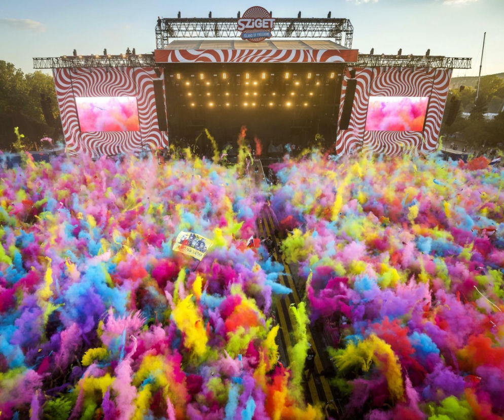 Tłum ludzi pod sceną Sziget Festival w chmurze kolorowych proszków holi. O dojeździe na festiwal przeczytasz w serwisie Eska.