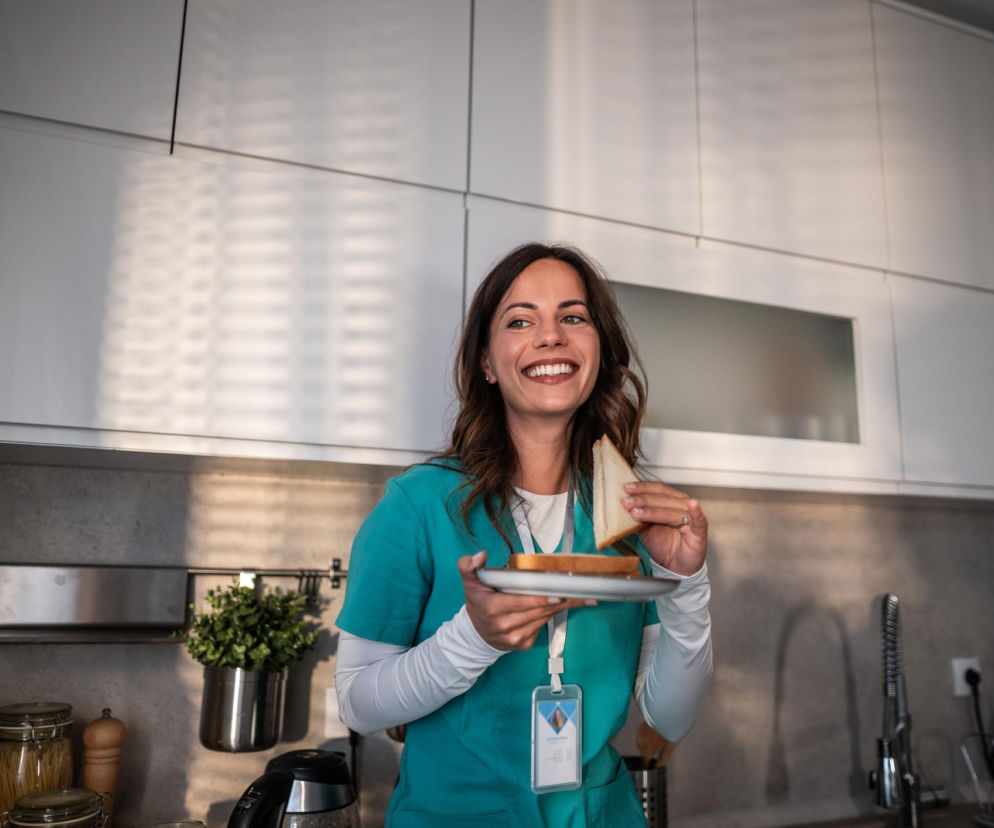 Uśmiechnięta kobieta w turkusowej bluzce medycznej trzyma talerzyk z kanapką i zjada kawałek, stojąc w jasnej kuchni. Ten obraz ilustruje błąd jedzenia na stojąco, który, jak czytać możesz na Poradnik Zdrowie, prowadzi do wzdęć i ciągłego głodu.