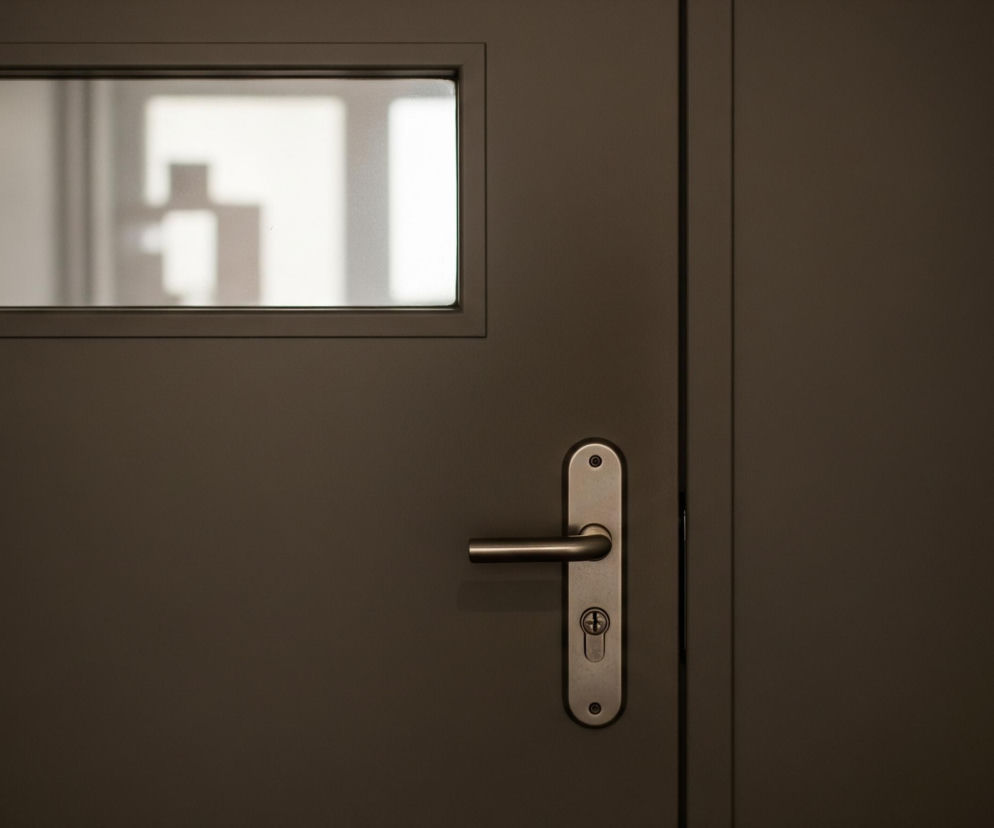 Brązowe drzwi z metalową klamką i zamkiem wypełniają lewą i środkową część kadru, a ich prawy skraj stapia się z ciemnością. W górnej, lewej części drzwi znajduje się prostokątne okienko z matowego szkła, przez które prześwituje rozmyty, biały kształt litery T na jasnym tle. Klamka i szyld zamka są w kolorze stali szczotkowanej, a na górze i na dole szyldu widać dwie śruby montażowe.