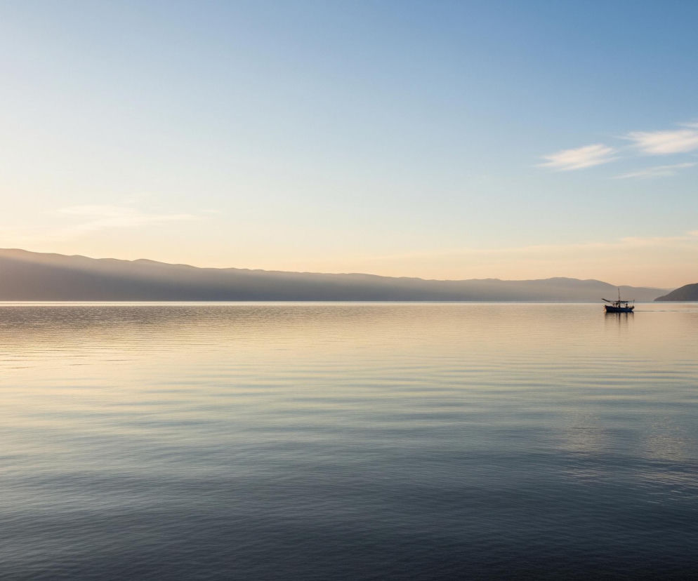 Na obrazie dominuje spokojny krajobraz jeziora w świetle zachodzącego słońca, z wyraźnym horyzontem oddzielającym niebo od wody. Horyzont przecina długie pasmo ciemnych, falistych gór lub wzgórz. Po prawej stronie w oddali, na powierzchni wody, pływa mała, ciemna łódź z widocznym masztem. Niebo stopniowo przechodzi od jasnego, złotego blasku po lewej, gdzie słońce jest najjaśniejsze, do pastelowego błękitu po prawej, z delikatnymi, białymi obłokami. Woda na pierwszym planie jest ciemnoniebieska, stopniowo rozjaśniając się ku horyzontowi do odcieni złocistych i jasnoniebieskich, z licznymi, drobnymi falami tworzącymi poziome linie na powierzchni.