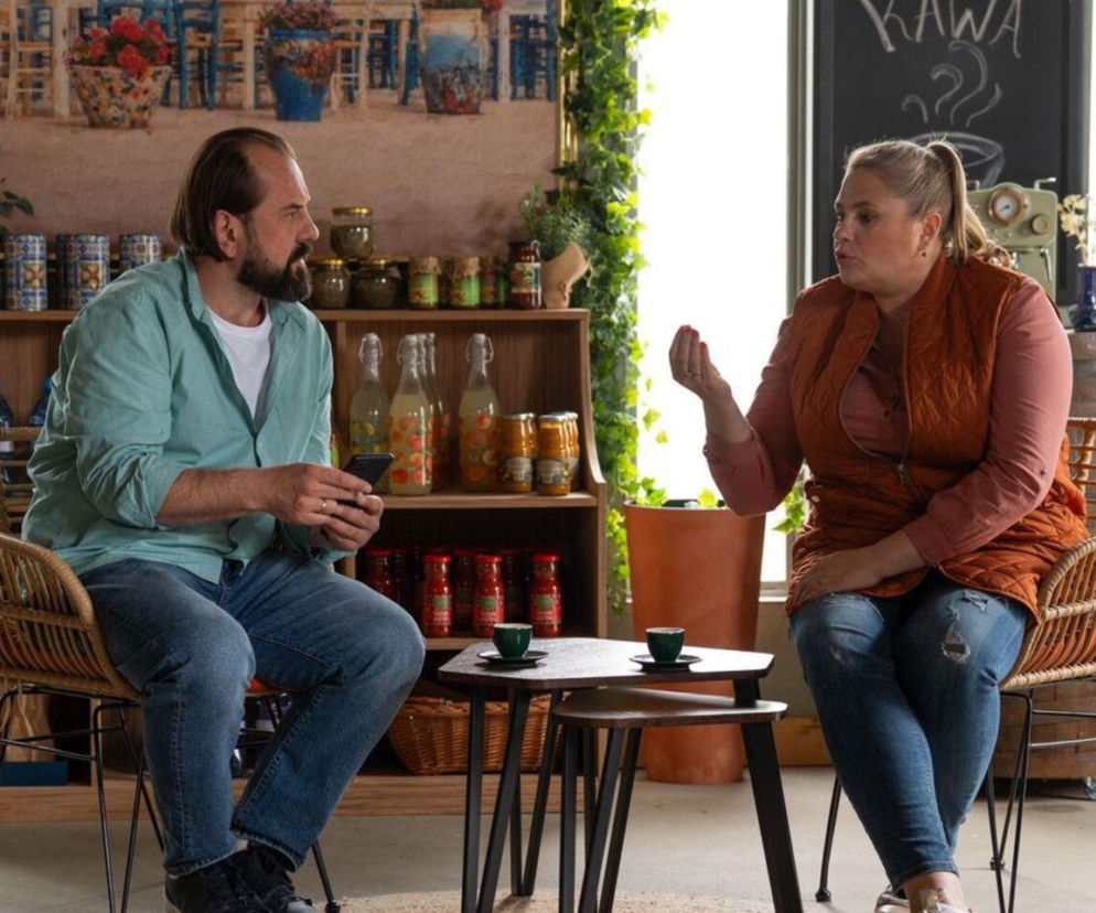 Aktorzy Elżbieta Romanowska jako Aldona i Jakub Wieczorek jako Borys z serialu Barwy szczęścia siedzą w rattanowych fotelach w pomieszczeniu imitującym kawiarnię. Aldona gestykuluje, Borys trzyma telefon, obok nich stoliki z filiżankami. Więcej o ich losach na Super Seriale.