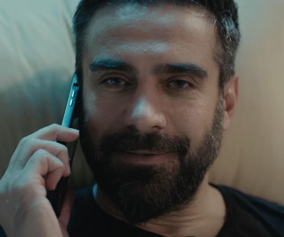 Uśmiechnięty mężczyzna z brodą rozmawia przez telefon. Mężczyzna leży, patrząc bezpośrednio w obiektyw, a w jego oczach widać blask. Zdjęcie ilustruje odcinek „Dziedzictwa”, o którym możesz przeczytać na Super Seriale.