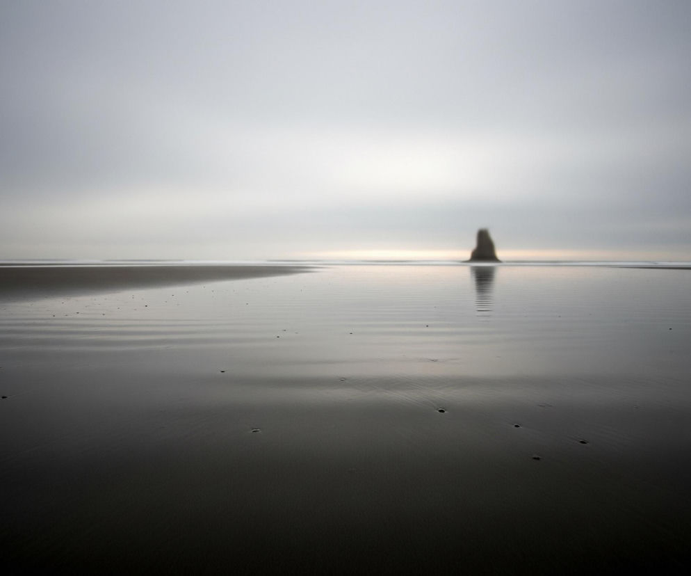 Płaska, rozległa plaża pokryta wilgotnym, ciemnym piaskiem rozciąga się w kierunku horyzontu. Piasek w pierwszym planie jest ciemnoszary, przechodząc w jaśniejszy odcień szarości bliżej linii morza, z pojedynczymi ciemnymi kamykami lub zagłębieniami rozrzuconymi na jego powierzchni. Na horyzoncie, na tle mglistego, jasnoszarego nieba, widoczna jest niewyraźna, ciemna formacja skalna o kształcie zbliżonym do stożka, z odbiciem w wodzie. Niebo jest jednolicie jasnoszare, lekko rozmyte, sugerując pochmurny dzień.