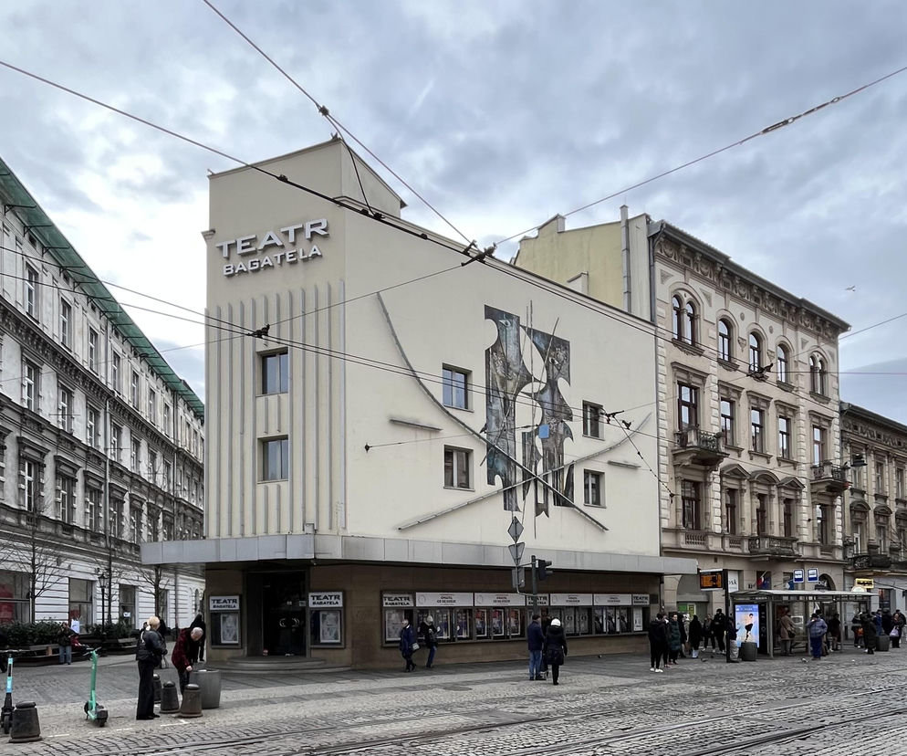 Teatr Bagatela w Krakowie z ceramicznymi kompozycjami Witolda Skulicza na elewacji, w otoczeniu ulicy z brukiem i tramwajowymi szynami. Z boku widać kamienicę z secesyjnymi zdobieniami. Przeczytaj o historii i przebudowach teatru na Architektura Murator Plus.