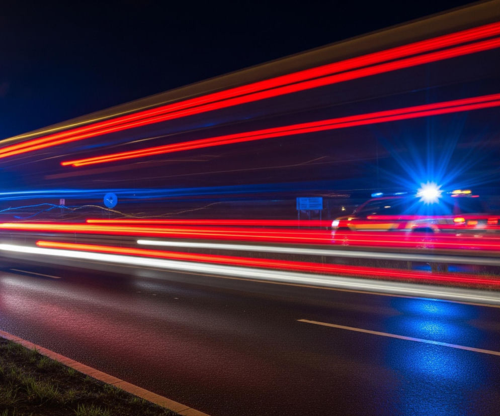 Nocna scena przedstawia autostradę oświetloną rozmazanymi smugami świateł pojazdów w ruchu. W dolnej części obrazu znajduje się ciemna nawierzchnia jezdni z białymi, rozmytymi pasami, odzwierciedlająca czerwone i niebieskie światła. Z lewej strony widoczne są intensywnie niebieskie i białe rozbłyski przednich świateł, a także wyraźne białe smugi. Na górze i po prawej stronie dominują długie, poziome smugi w kolorze czerwonym, z kilkoma żółtymi i niebieskimi akcentami, tworzące dynamiczne linie na tle ciemnego nieba.