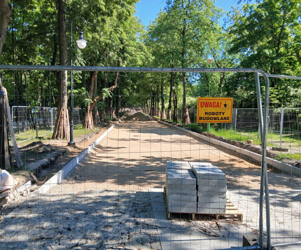 Aleja w Parku Planty w Białymstoku otoczona metalowym ogrodzeniem budowlanym i drzewami. Na pierwszym planie stosy kostki brukowej i znak UWAGA! ROBOTY BUDOWLANE, symbolizujące trwającą przebudowę, o której można przeczytać na Architektura Murator Plus.