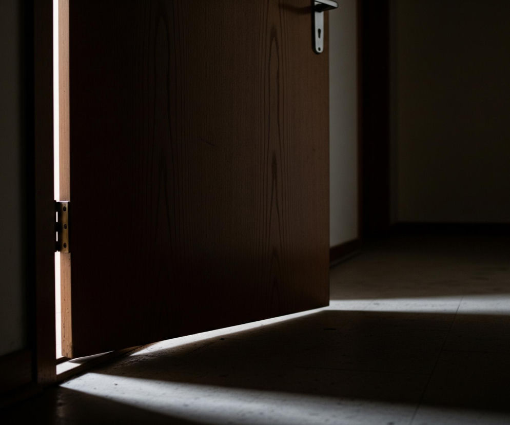 Uchylone drewniane drzwi z metalową klamką w kształcie litery T tworzą pionową, rozświetloną szczelinę po lewej stronie. Intensywne światło zza drzwi pada na podłogę, tworząc ukośny, jasny pas, który przechodzi przez ciemne pomieszczenie. Reszta pokoju, w tym ściany i podłoga, pozostaje w głębokim cieniu, podkreślając kontrast między jasnością a mrokiem.