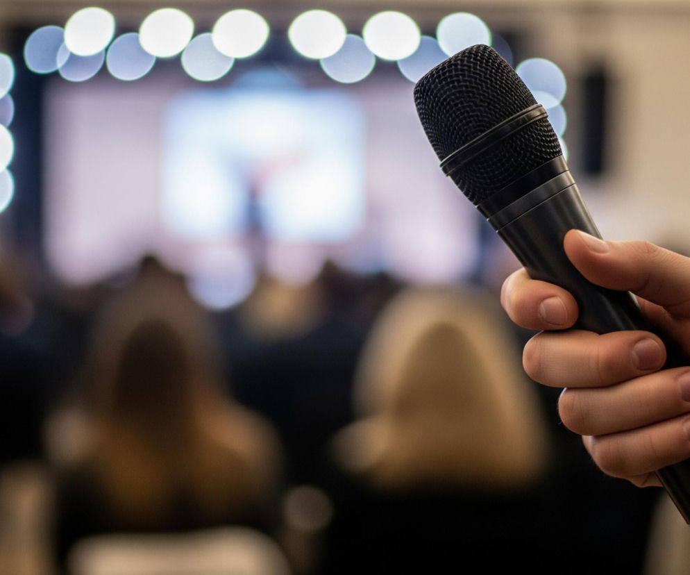 Prawa dłoń, ubrana w ciemną marynarkę z białą koszulą, trzyma czarny mikrofon, który zajmuje prawą stronę pierwszego planu. Tło jest rozmyte, ukazując salę wypełnioną niewyraźnymi sylwetkami ludzi w ciemnych ubraniach. W głębi widoczny jest ekran projekcyjny z niewyraźnym obrazem i jaskrawe, okrągłe punkty świetlne, tworzące efekt bokeh.