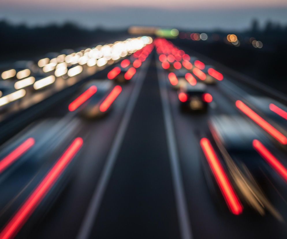 Autostradę w nocy, z rozmytymi smugami świateł pojazdów. Dwie główne jezdnie biegną równolegle przez centrum kadru, rozdzielone niewyraźnym pasem zieleni. Lewa jezdnia jest wypełniona białymi i żółtymi smugami świateł przednich samochodów jadących w kierunku oddalania się od widza. Prawa jezdnia jest zajęta przez czerwone smugi świateł tylnych pojazdów poruszających się w kierunku zbliżania się do widza. Tło jest ciemne i niewyraźne, z zarysem drzew na horyzoncie i bladym, fioletowo-różowym niebem.
