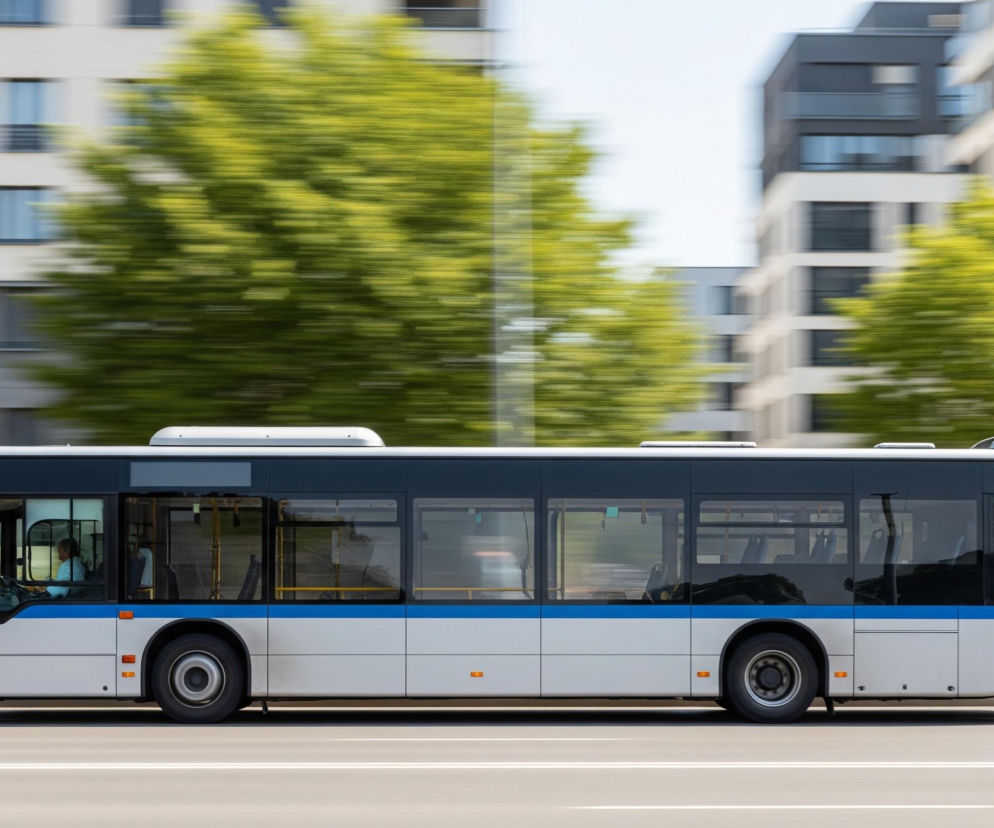 Na obrazie dominuje widok boczny autobusu jadącego w prawo, z efektem rozmycia tła sugerującym ruch. Autobus jest biało-szary z poziomym niebieskim paskiem biegnącym przez całą długość pojazdu, tuż pod oknami. Wewnątrz autobusu widać siedzenia pasażerskie, a za przednią szybą widoczna jest postać kierowcy. Tło składa się z rozmazanych zielonych drzew i białych budynków z ciemnymi oknami, co wzmacnia wrażenie prędkości.