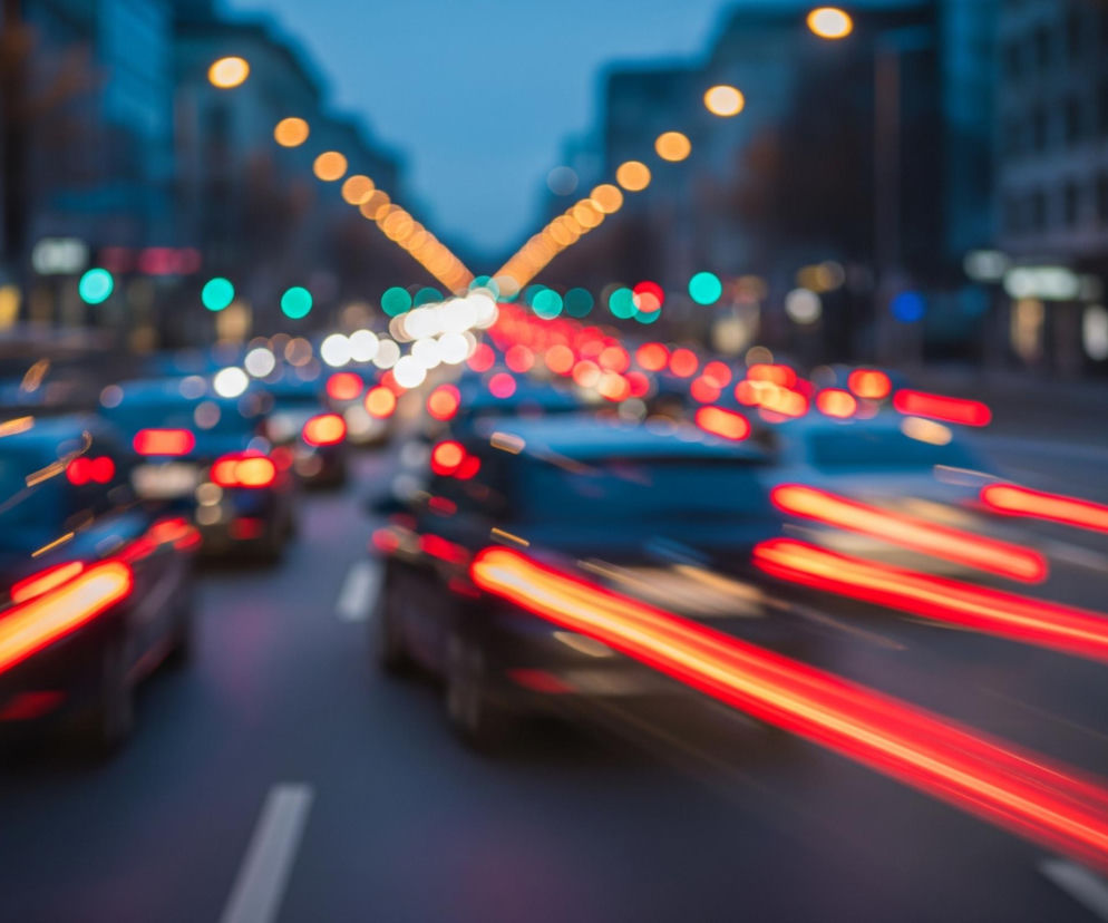 Ulica miejska wypełniona jest jadącymi samochodami, uwiecznionymi techniką długiej ekspozycji. Czerwone smugi świateł tylnych pojazdów dominują w dolnej części obrazu, rozciągając się dynamicznie w prawą i lewą stronę, tworząc wrażenie ruchu i prędkości. Na jezdni widoczne są również białe, przerywane pasy. W tle, powyżej samochodów, rozmywają się jaskrawe, okrągłe światła uliczne w kolorach pomarańczowym, białym i zielonym, tworzące efekt bokeh. Odcienie szarości i granatu dominują w krajobrazie miejskim, z niewyraźnymi zarysami budynków po obu stronach ulicy.