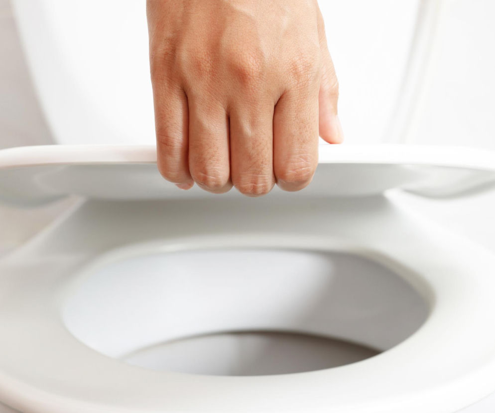 Dłoń osoby podnoszącej białą klapę sedesu w sterylnej, jasnej toalecie, symbolizująca problem z parurezą, czyli zespołem nieśmiałego pęcherza. Przeczytaj o przyczynach i trikach na blokadę w publicznej toalecie na Poradnik Zdrowie.