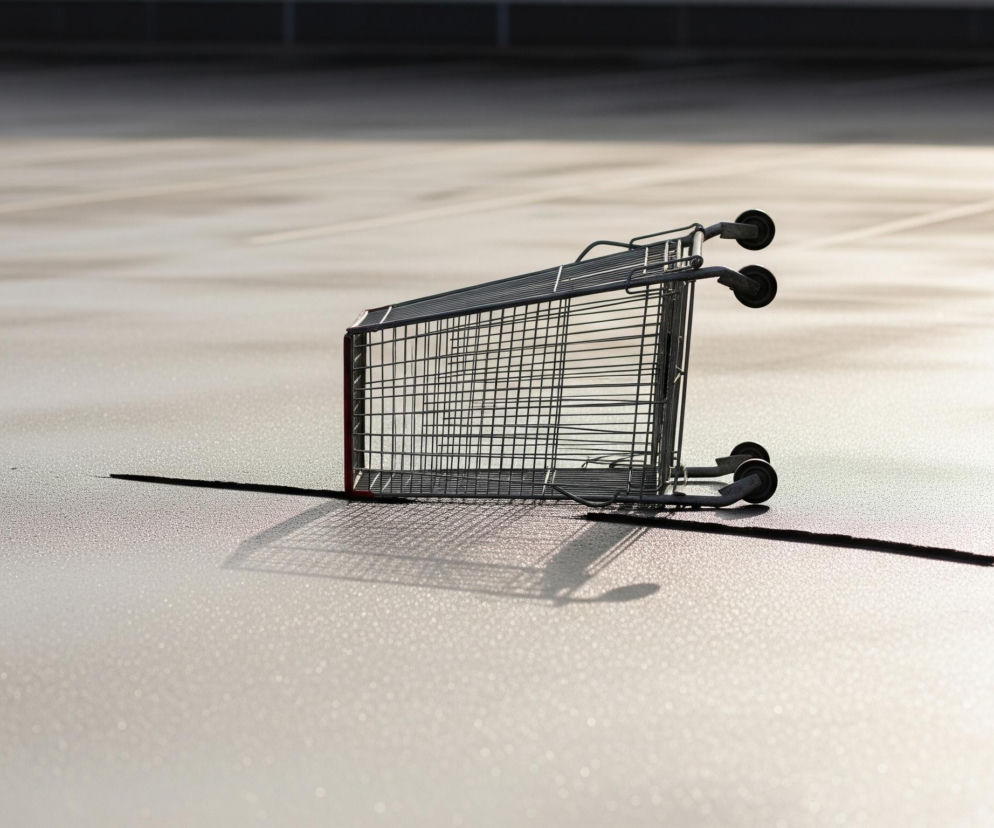 Na obrazie centralnie leży przewrócony, metalowy wózek na zakupy, składający się z siatkowych ścianek i czterech kółek, z czego dwa są w górze. Wózek rzuca długi, wyraźny cień na jasną, równą powierzchnię, która wydaje się być parkingiem lub placem, charakteryzującym się delikatną, ziarnistą teksturą i błyszczącymi punktami. W tle widać ciemniejszą, rozmytą przestrzeń, sugerującą ścianę lub dalsze zabudowania. Całość jest oświetlona z prawej strony, co tworzy silne kontrasty światła i cienia.