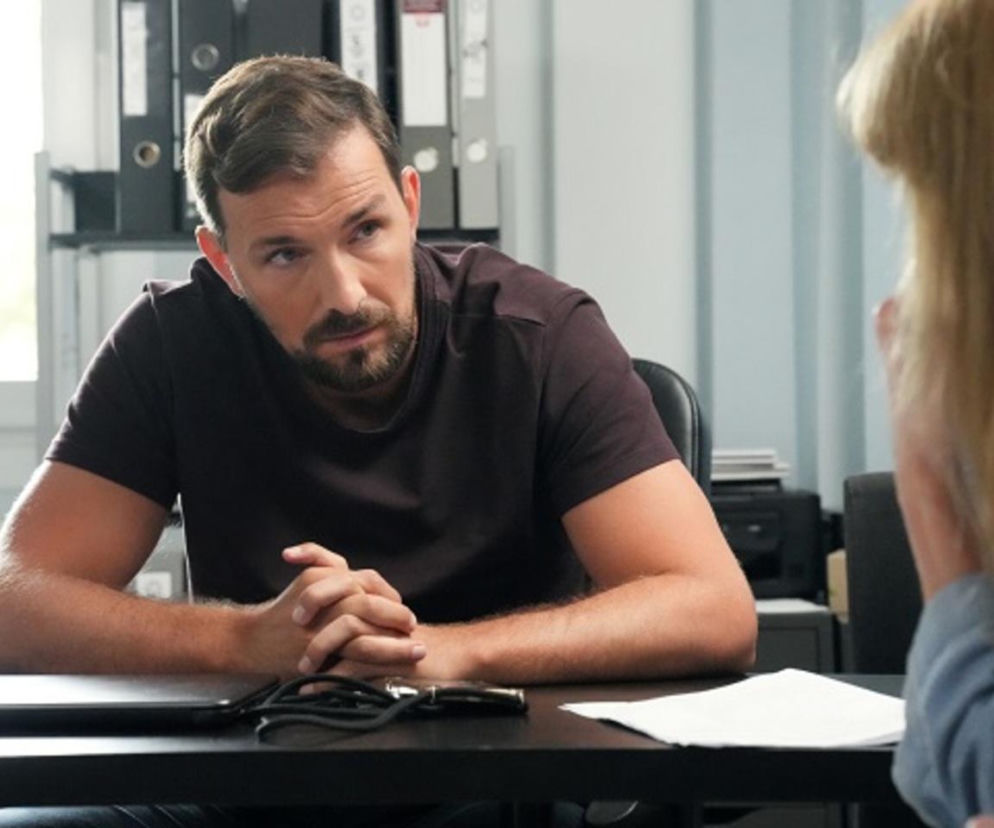 Marcin Kodur (Oskar Stoczyński) z serialu Barwy szczęścia w ciemnej koszulce, z brodą, siedzi przy stole z zaciśniętymi dłońmi, rozmawia z niewidoczną kobietą podczas przesłuchania Agnieszki. Więcej o wątkach przeczytasz na Super Seriale.