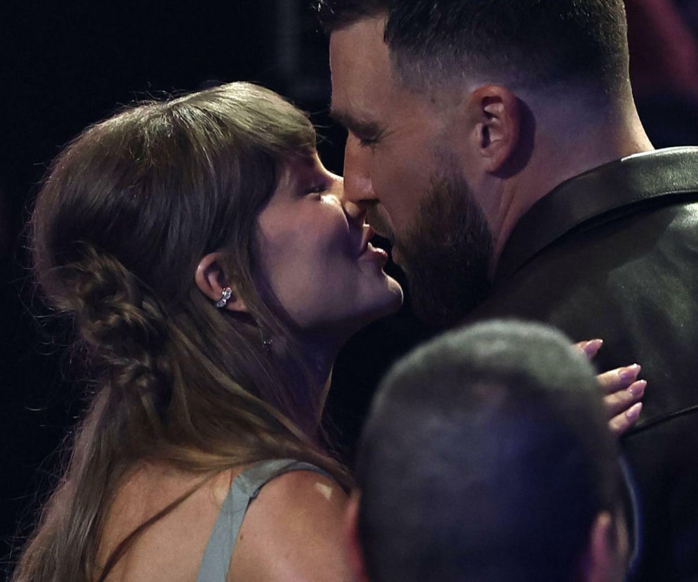 Taylor Swift z Travisem Kelce całują się na tle ciemnego, rozmytego tła. Widać bliską relację pary, o której ślubie można przeczytać na naszym portalu. Ona w jasnym topie z warkoczem i kolczykiem, on w ciemnej kurtce.