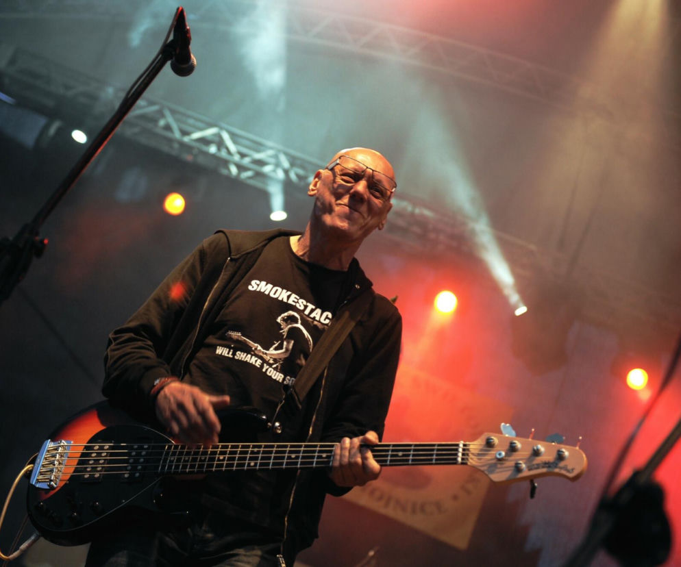 Andrzej „Długi” Laskowski w okularach, grający na gitarze basowej na scenie, w tle reflektory i mikrofon. Muzyk ubrany w czarną koszulkę i bluzę. Więcej o zmarłym basiście przeczytasz na naszym portalu.