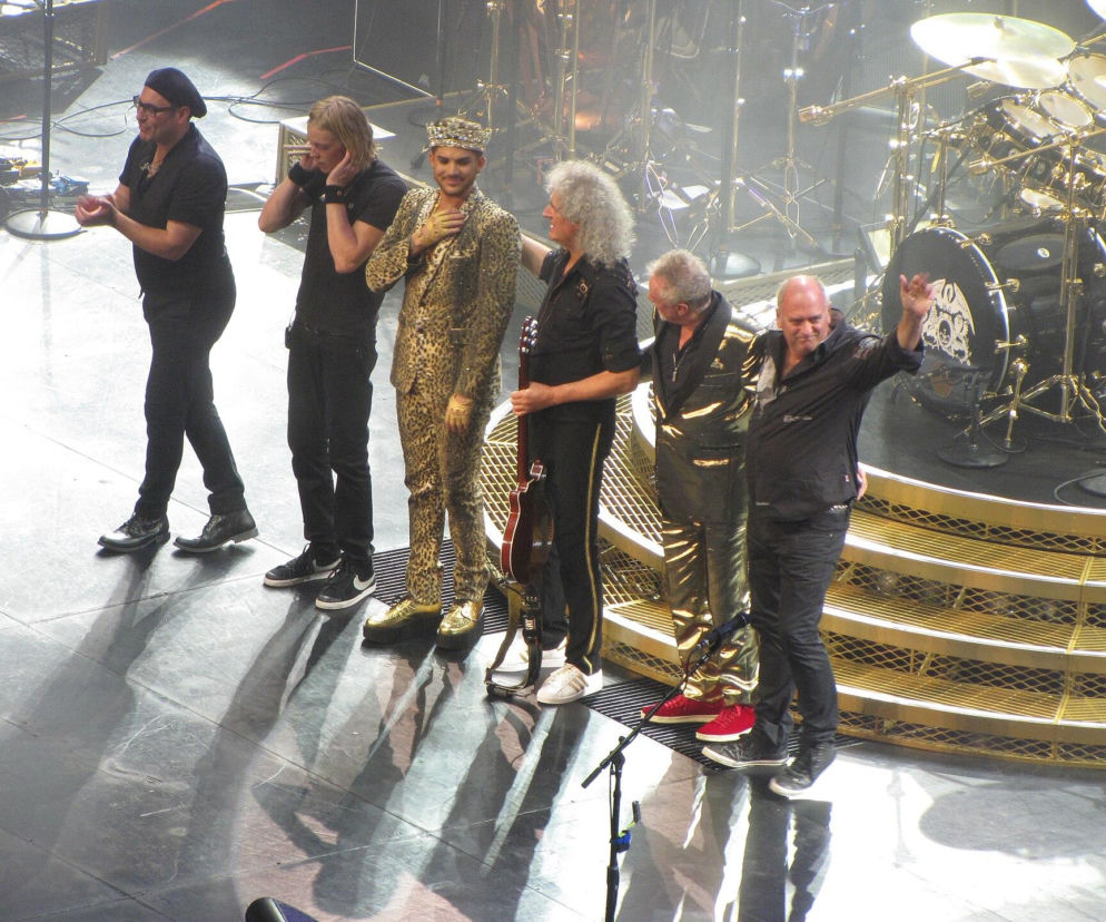 Zespół Queen + Adam Lambert na scenie podczas koncertu. Adam Lambert w leopardowym garniturze i koronie, obok niego Brian May z gitarą, Roger Taylor w złotym kombinezonie i dwóch innych muzyków. Więcej o ich dalszych losach przeczytasz na naszym portalu.