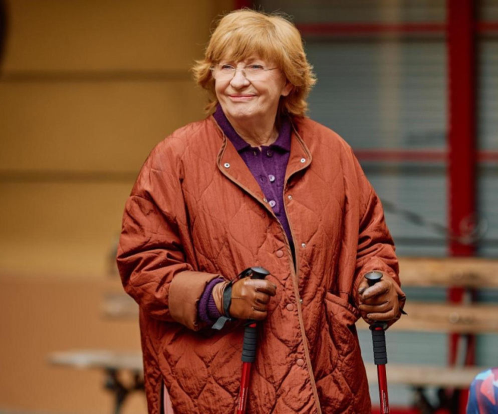 Uśmiechnięta Krystyna z Na Wspólnej w brązowej kurtce trzyma kijki do nordic walking. O jej losach czytaj w SE Superseriale.