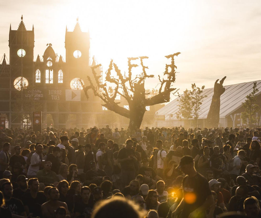 Tłum ludzi na festiwalu Hellfest w Clisson we Francji, w tle scenografia w kształcie zamku, drzewa z napisem HELLFEST i uniesionej pięści, wszystko skąpane w złotym świetle zachodzącego słońca. O festiwalu Hellfest 2026 i koncertach przeczytasz więcej na naszym portalu.