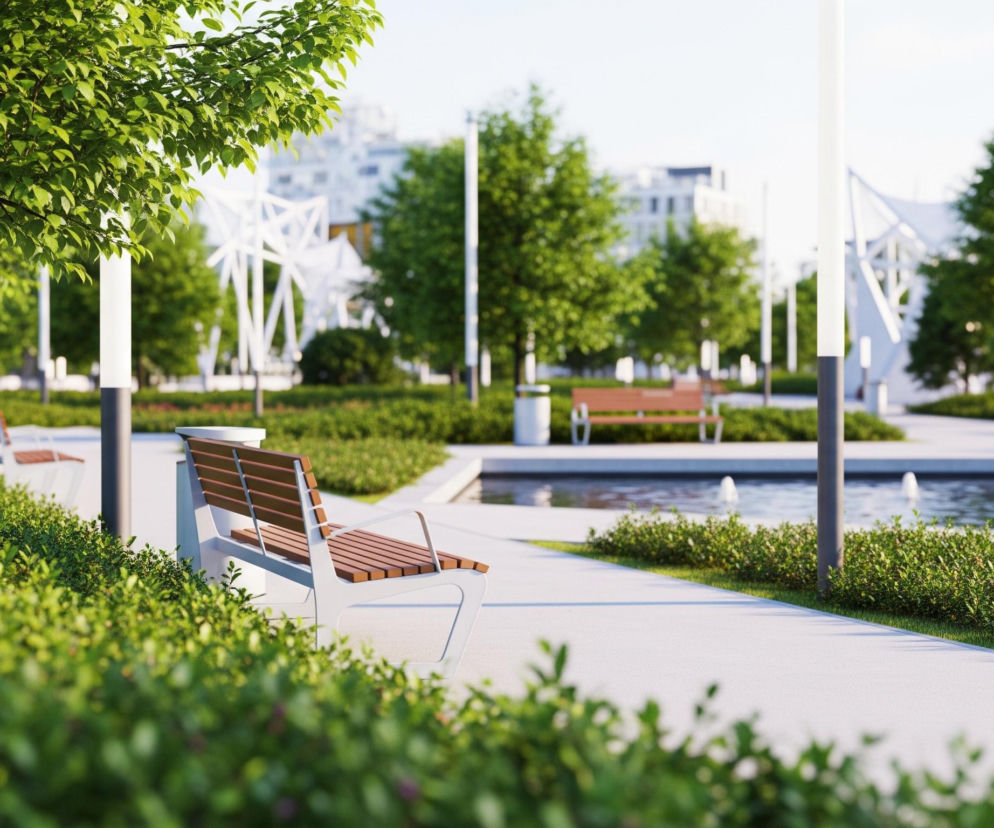 Na pierwszym planie, z prawej strony, stoi nowoczesna ławka z jasnoszarym metalowym stelażem i drewnianymi, brązowymi deskami siedzenia i oparcia, otoczona gęstymi, zielonymi krzewami. Dalej, po lewej stronie, widoczne są pnie drzew z jasnozielonymi liśćmi, a pod nimi rozciągają się żywe, zielone zarośla. W centrum obrazu znajduje się szeroka, jasnoszara ścieżka spacerowa, która prowadzi w głąb kadru, z prawej strony ścieżki rozciąga się prostokątny zbiornik wodny z trzema fontannami, z których tryskają białe strumienie wody. W tle majaczą niewyraźne białe konstrukcje o geometrycznych kształtach oraz fragmenty budynków w podobnej kolorystyce, a pomiędzy nimi drzewa o intensywnie zielonych koronach.