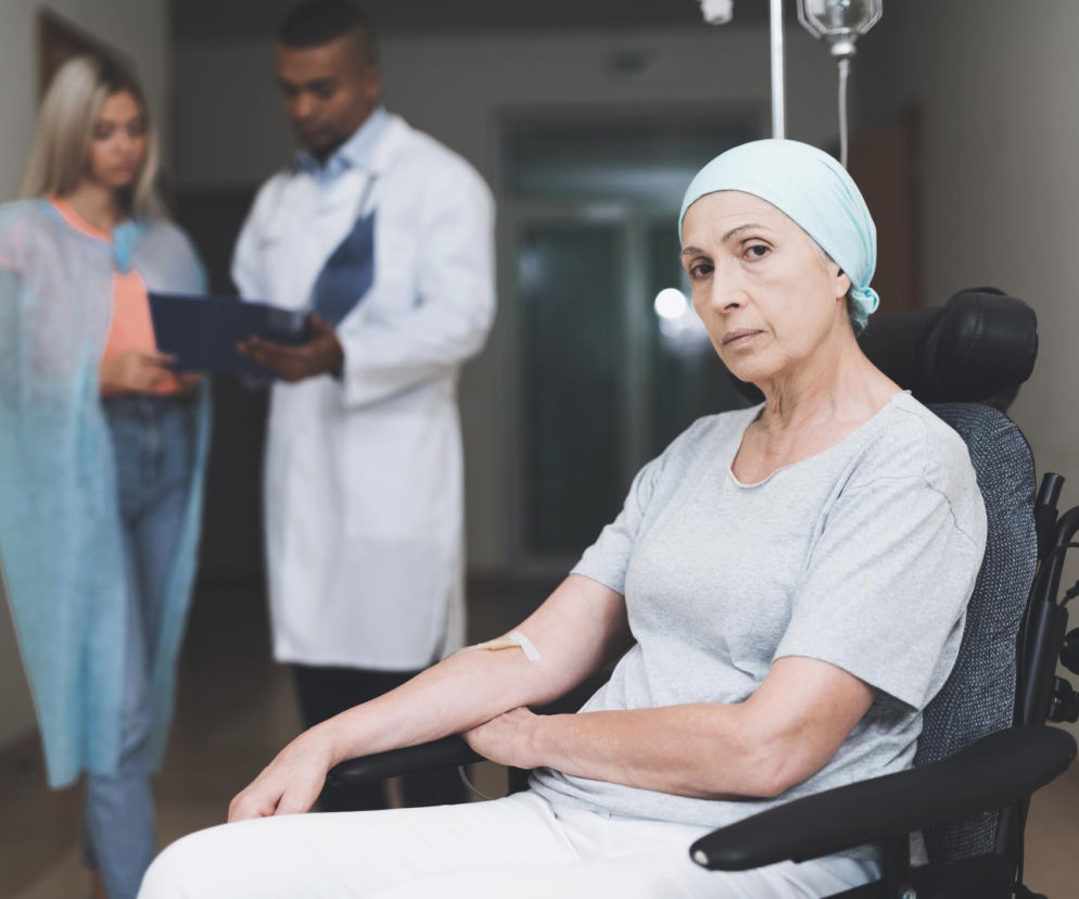 Starsza kobieta w jasnej chuście na głowie i szarej koszulce siedząca na wózku inwalidzkim z wkłuciem na ramieniu, w tle rozmazany lekarz i osoba towarzysząca. Obraz symbolizuje walkę z nowotworem i wsparcie medyczne, o czym więcej przeczytasz na Poradnik Zdrowie.