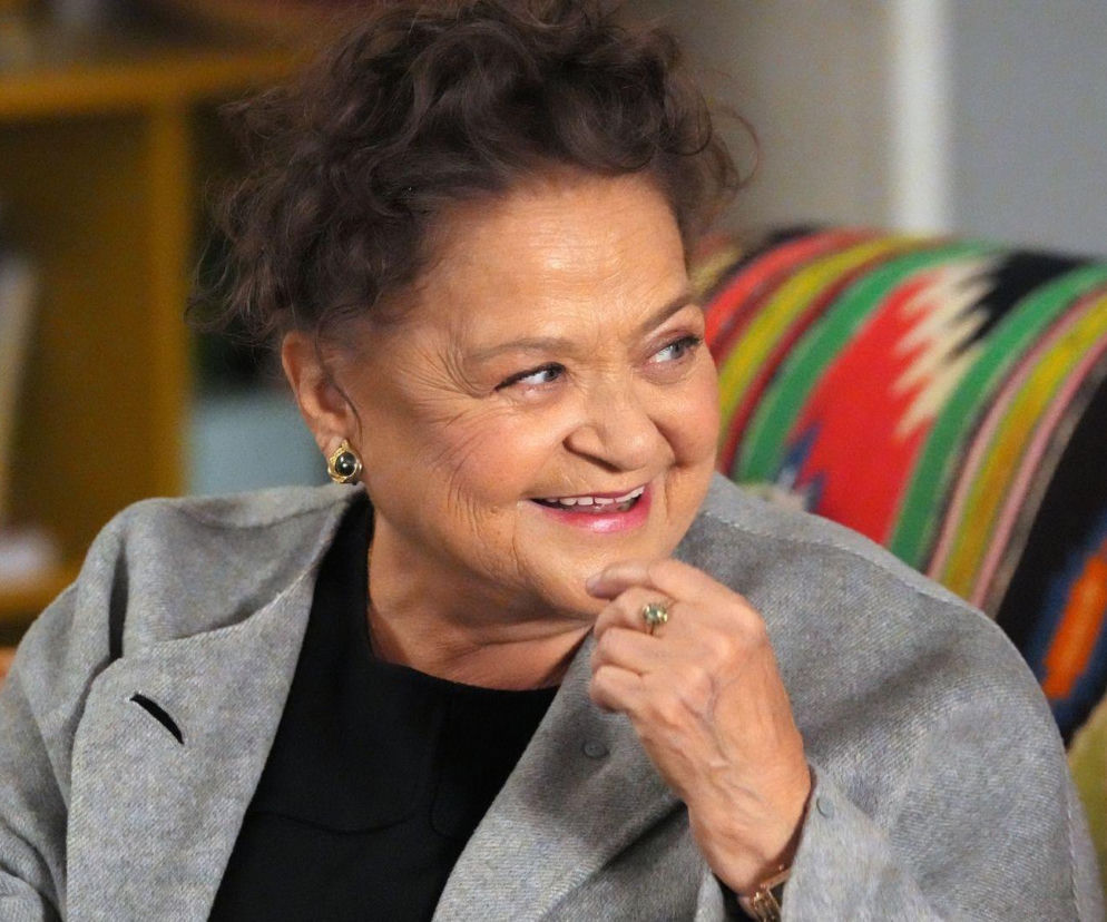 Uśmiechnięta kobieta w średnim wieku, Lucyna Zwierzchowska, w jasnej marynarce, z kręconymi włosami i kolczykami, opiera dłoń na brodzie, patrząc w bok. W tle widoczna jest kolorowa poduszka i żółty regał. O wydarzeniach z jej udziałem, dowiesz się więcej na Super Seriale.