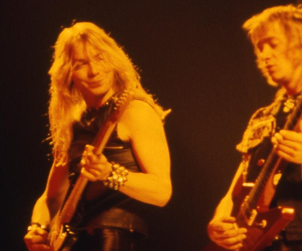 Muzycy Iron Maiden, Janick Gers z długimi blond włosami i skórzanej kamizelce z ćwiekami, grający na gitarze, oraz drugi, prawdopodobnie Dave Murray, w kamizelce w panterkę. Obraz oddaje energię koncertu zespołu, o którym więcej przeczytasz na naszym portalu.