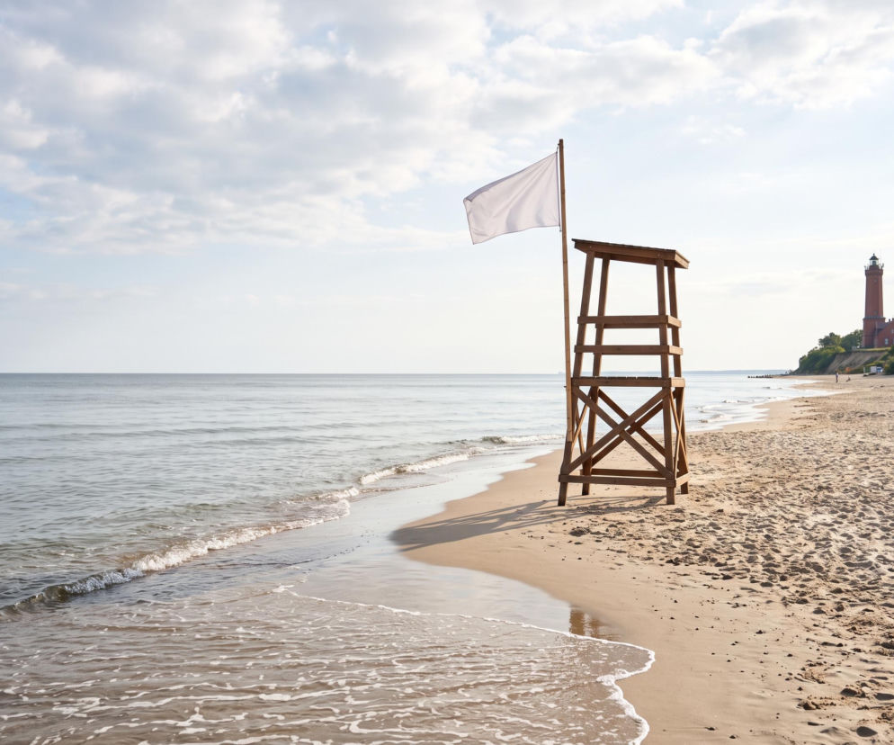 Pusta plaża w Niechorzu z białą flagą na wieży ratowniczej i latarnią morską w tle. O pogodzie przeczytasz na SE Szczecin.