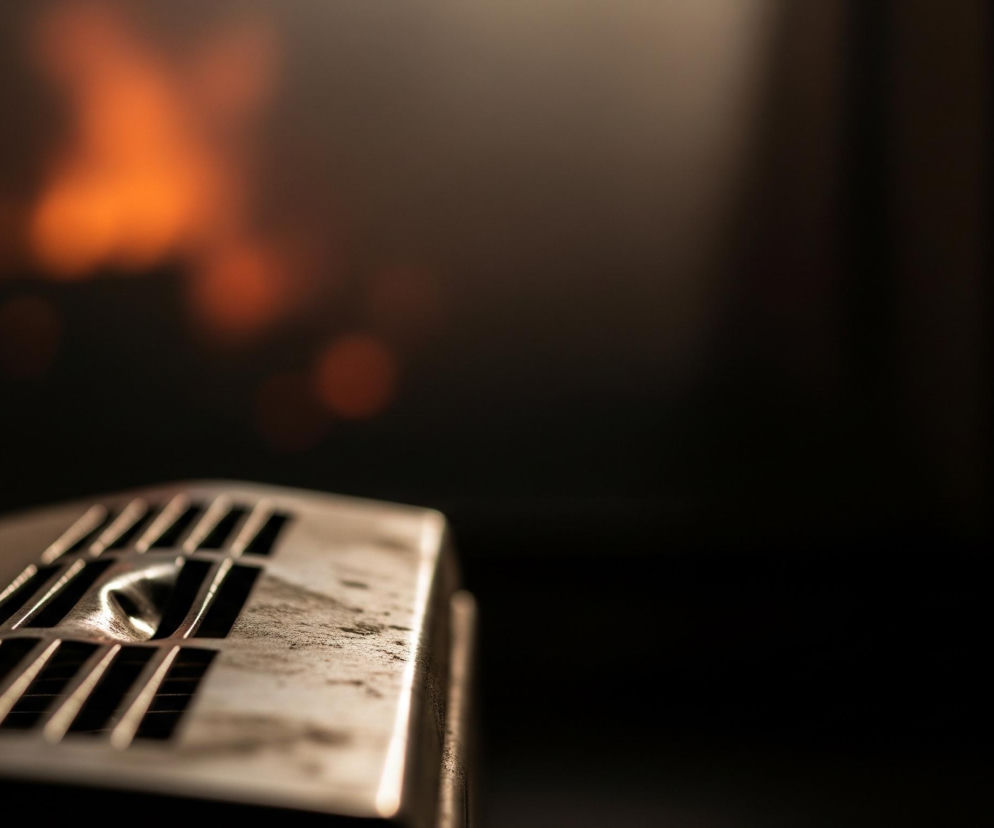 Na pierwszym planie, w dolnej części kadru, znajduje się ostro zogniskowany, metalowy element, przypominający kratkę wentylacyjną lub część urządzenia, o barwie srebrzystej z widocznymi przebarwieniami i osadami. Posiada on podłużne szczeliny oraz jeden okrągły otwór z wypukłym pierścieniem. Tło jest rozmyte, z niewyraźnymi, pomarańczowo-czerwonymi plamami światła, sugerującymi ogień lub jego odbicie, na ciemnym, niemal czarnym tle.