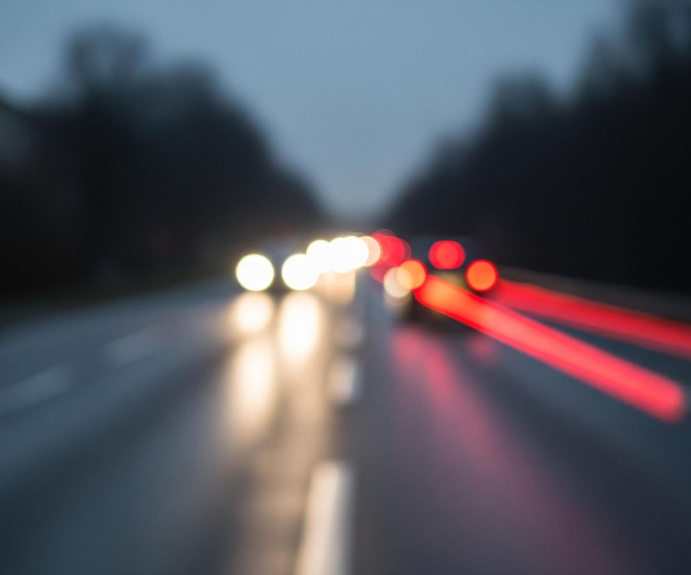 Rozmazana scena przedstawia widok z perspektywy jadącego pojazdu, skupiając się na drodze o zmierzchu lub w nocy. Na środku jezdni widoczne są rozmyte białe światła nadjeżdżających samochodów, tworzące jasne, okrągłe plamy. Po prawej stronie drogi widać wydłużone, czerwone smugi świateł pozycyjnych lub tylnych lamp innych pojazdów, sugerujące ruch w przeciwnym kierunku lub oddalanie się. Droga ma ciemną, wilgotną powierzchnię, odbijającą częściowo światła, a po jej bokach rozciągają się niewyraźne, ciemne sylwetki drzew lub budynków.