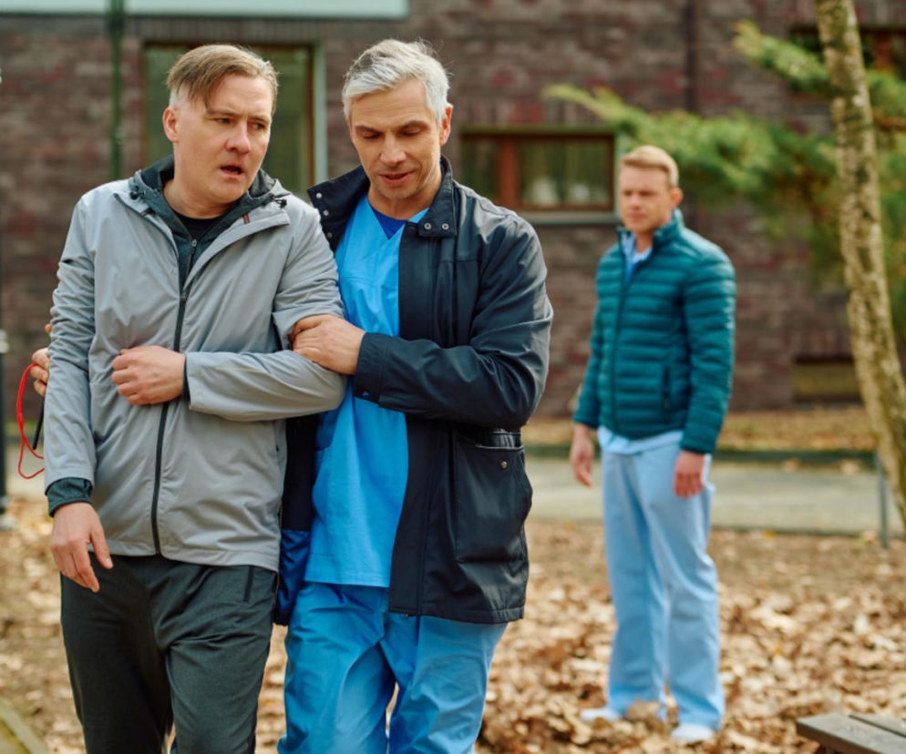 Mężczyzna w szarej kurtce sportowej, Tomasz Adamski (Tomasz Piątkowski), jest podtrzymywany przez kardiochirurga Grega (Michał Sitarski) w niebieskim stroju medycznym, idących przez park po morderczym treningu. W tle stoi kolejny lekarz. Więcej o ich losach przeczytasz na naszym portalu.