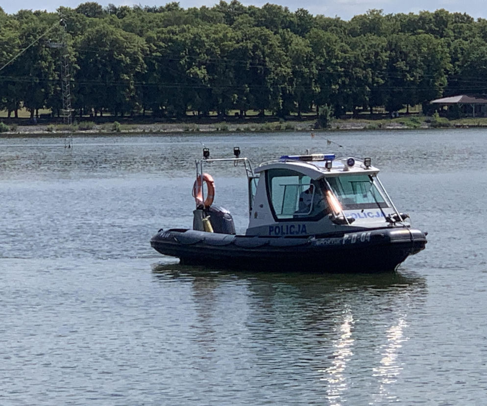 Łódź patrolowa policji z napisem POLICJA na burcie, wyposażona w światła ostrzegawcze i koło ratunkowe, pływająca po jeziorze, z zielonym, zalesionym brzegiem w tle. Zdjęcie ilustruje akcję Bezpieczne Wakacje w województwie lubelskim, o której przeczytasz więcej na naszym portalu.