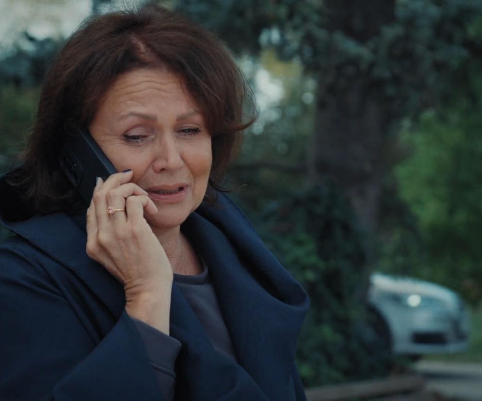 Małgorzata Pieńkowska jako Marysia Rogowska z M jak miłość, smutna kobieta w ciemnym płaszczu rozmawia przez telefon, trzymając smartfon przy uchu, na jej palcu widać pierścionek z różowym oczkiem. W tle rozmyta zieleń drzew i fragment samochodu. Więcej o losach bohaterów na Super Seriale.