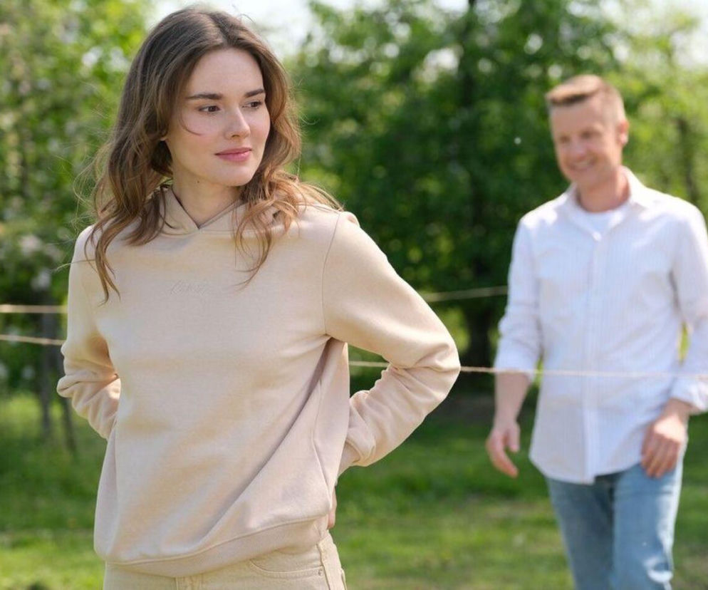 Młoda kobieta, Dominika Kachlik jako Franka z M jak miłość, ubrana w beżową bluzę z kapturem, z rękami na biodrach, patrzy w bok na tle zieleni. W tle widać uśmiechniętego mężczyznę, Pawła Zduńskiego, w białej koszuli i jeansach. To kadr z serialu, o którym przeczytasz więcej na Super Seriale.