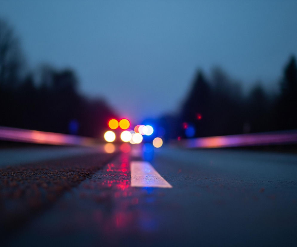 Droga z wilgotną, ciemną nawierzchnią rozciąga się w głąb obrazu, zajmując dolną połowę kadru. Na niej widoczna jest wyraźna, biała linia przerywana, której część w przedniej części obrazu jest ostra, a w dalszej rozmyta. Po lewej i prawej stronie drogi widać rozmyte, ciemne sylwetki drzew na tle bladoniebieskiego, zmierzchowego nieba.

W oddali, na horyzoncie, znajdują się jasne, rozmyte światła: czerwone, pomarańczowe i niebieskie, przypominające światła pojazdów ratunkowych lub ostrzegawczych. Ich odbicia, w postaci czerwonych i niebieskich smug, są widoczne na mokrej nawierzchni drogi. W górnych częściach lewej i prawej krawędzi obrazu widoczne są również rozmyte, podłużne, fioletowo-różowe smugi światła.