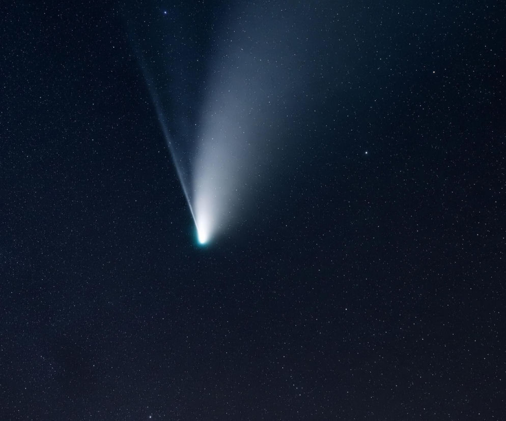 Na ciemnoniebieskim niebie, usianym niezliczonymi drobnymi, białymi punktami gwiazd, dominuje jasny obiekt przypominający kometę. Obiekt ten posiada wyraźne, turkusowo-niebieskie jądro w dolnej części obrazu, z którego rozciąga się szeroki, biały i rozmyty ogon, skierowany ukośnie w prawo i w górę. Ogon rozprasza się stopniowo, stając się coraz mniej intensywny w miarę oddalania się od jądra. Niebo jest ciemniejsze w lewym dolnym rogu i jaśniejsze w prawym górnym, gdzie ogon komety wydaje się jeszcze bardziej rozświetlać otoczenie.