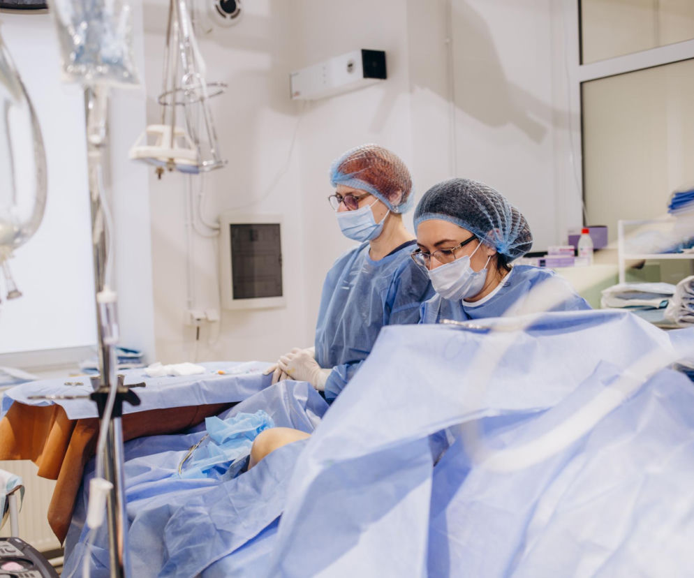 Dwie kobiety w strojach medycznych, czepkach i maseczkach, pochylają się nad pacjentem przykrytym niebieską tkaniną chirurgiczną, sugerującym zabieg operacyjny. Obraz przedstawia scenę z sali operacyjnej. O błędach medycznych przeczytasz na portalu Poradnik Zdrowie.