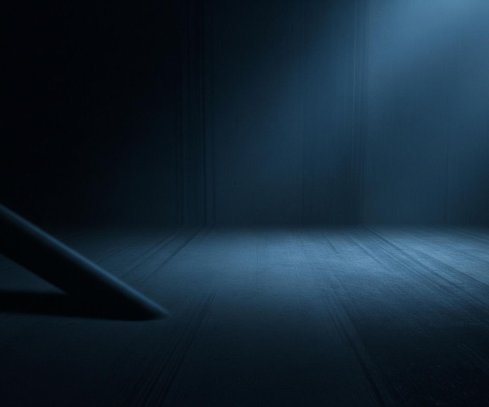 Ciemne, puste pomieszczenie lub scenę utrzymaną w odcieniach głębokiego granatu i błękitu. Światło pada z góry, z prawej strony, oświetlając fragment tylnej ściany oraz podłogę, tworząc jasny punkt na ich styku. Lewa część kadru jest znacznie ciemniejsza, z widocznym, ukośnym, ciemnym obiektem, przypominającym walec lub rurę, leżącym na podłodze i rzucającym długi cień w lewo. Tekstura podłogi i ściany wydaje się szorstka, z delikatnymi pionowymi i poziomymi liniami, które podkreślają wrażenie przestrzenności.