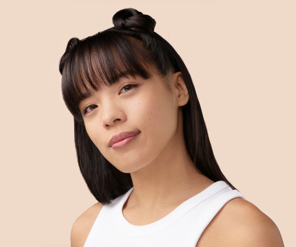 Młoda kobieta z ciemnymi włosami, ułożonymi w dwa koki i z grzywką, z widocznymi na skórze niewielkimi niedoskonałościami, co symbolizuje problem trądziku. Twarz modelki jest skierowana lekko w bok z delikatnym uśmiechem, prezentuje pewność siebie w kontekście nowoczesnej pielęgnacji cery trądzikowej, o której możesz przeczytać na Poradnik Zdrowie.