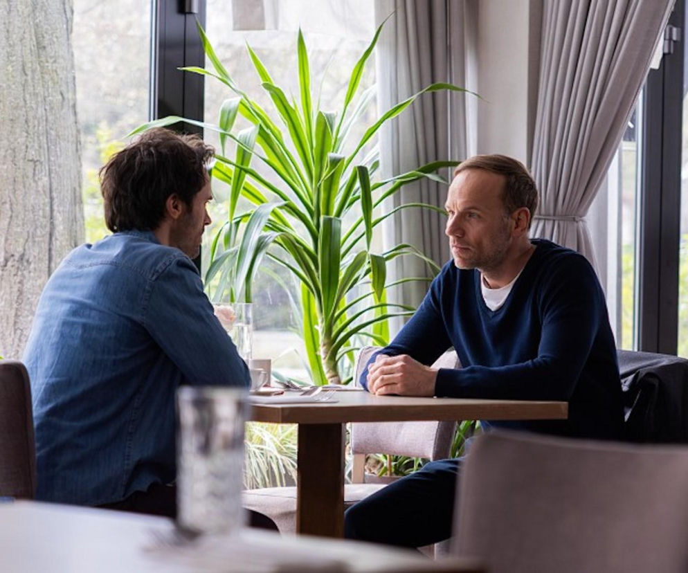 Dwóch mężczyzn, Maciej Mikołajczyk jako Janek i Mirosław Kropielnicki, siedzi przy stoliku w kawiarni, prowadząc poważną rozmowę. Widok przez okno ukazuje roślinność, sugerując naturalne otoczenie. Emocje związane z serialem Pierwsza miłość i jego finałowym odcinkiem znajdziesz na Super Seriale.