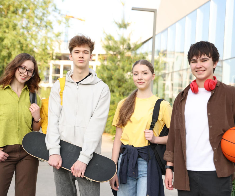 Uśmiechnięci nastolatkowie z plecakami, deskorolką i piłką do koszykówki stoją na zewnątrz budynku. Reprezentują młodych ludzi, których preferencje dotyczące napojów analizuje Super Biznes.
