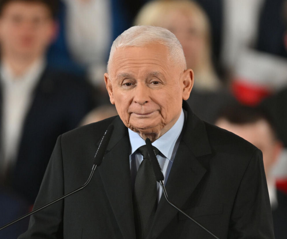 Jarosław Kaczyński, prezes PiS, przemawia za mównicą z dwoma mikrofonami, ubrany w czarny garnitur i krawat oraz błękitną koszulę. Na naszym portalu możesz przeczytać o jego problemach w szkole.