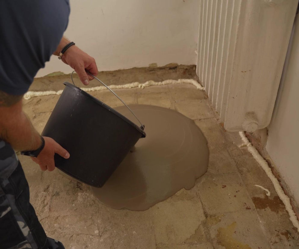 Osoba wylewająca masę samopoziomującą z czarnego wiadra na podłogę. O pracach remontowych przeczytasz na Muratordom.