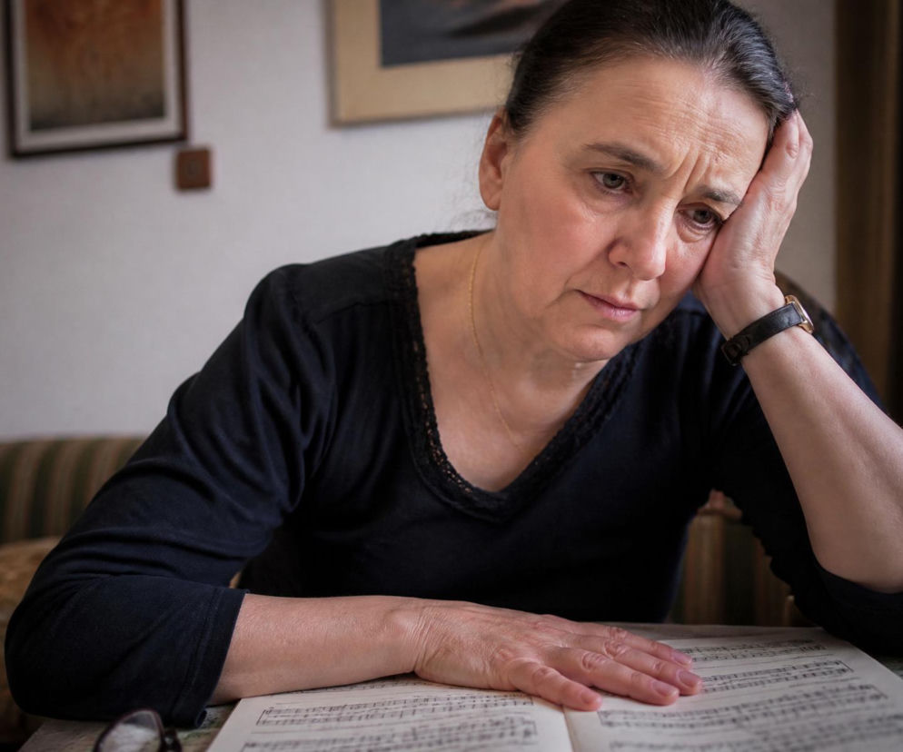 Kobieta w średnim wieku z zatroskaną miną, w czarnej bluzce z dekoltem i koronką, opiera głowę na dłoni, siedząc przy stole z otwartym zeszytem nutowym, co symbolizuje jej zmartwienie konfliktem o wychowanie wnuka, o czym przeczytasz na naszym portalu.