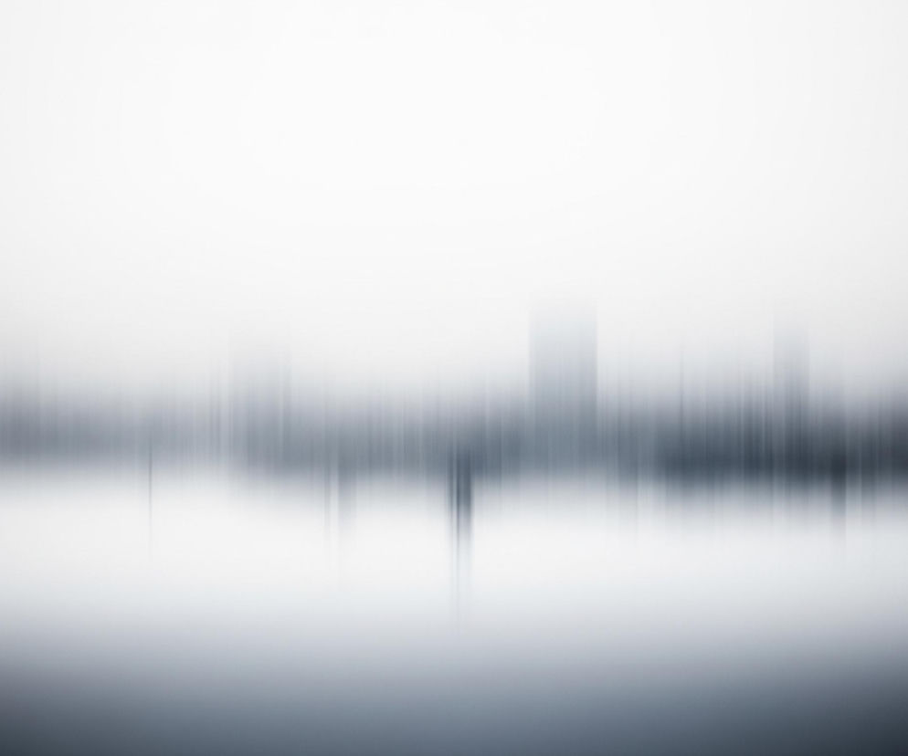 Pozioma kompozycja charakteryzuje się rozmyciem ruchu, tworząc abstrakcyjny krajobraz. Górna część obrazu jest jasnoszara, przechodząca w ciemniejszy, niebieskawo-szary odcień w dolnej połowie. W środkowej części widoczne są pionowe, ciemne smugi, sugerujące zamazany horyzont z odległymi obiektami lub drzewami, odbijającymi się w powierzchni wody poniżej. Całość emanuje spokojną, eteryczną atmosferą.
