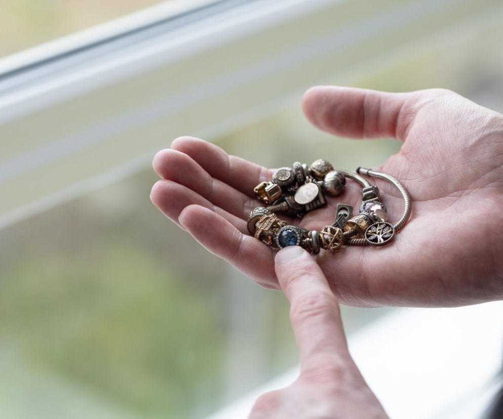 Osoba trzymająca srebrną bransoletkę typu Pandora w dłoni, wskazując palcem na jeden z ciemniejących charmsów. Tło rozmyte, widoczne okno i zieleń za nim. W artykule na Poradnik Zdrowie znajdziesz informacje, dlaczego srebrna biżuteria ciemnieje.