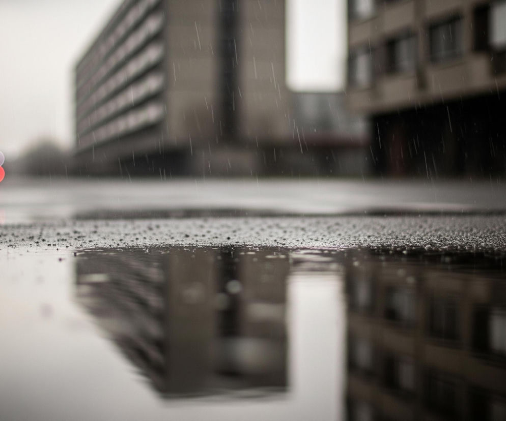 Asfaltowa powierzchnia z licznymi drobnymi kroplami wody w pierwszym planie odbija zniekształcony obraz otoczenia. Na mokrej nawierzchni widoczne są odbicia budynków, koloru szarego i brązowego, z ciemnymi oknami, rozciągających się w tle. Na lewo, w oddali, zamglone światło drogowe świeci na czerwono, poniżej niewyraźnego białego światła. Delikatne, białe smugi deszczu przecinają powietrze na całej szerokości kadru, a cała scena utrzymana jest w stonowanych odcieniach szarości i brązu, z rozmyciem tła.